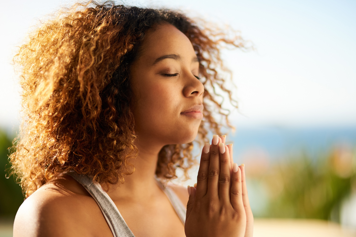 A closeup image of a woman with her eyes closed and her hands pressed together, she appears to be meditating. She looks like she is finding inner peace.