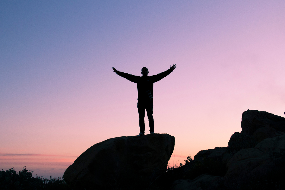 A sunset or sunrise image. We see the silhouette of a person with their back to us and their arms spread out.