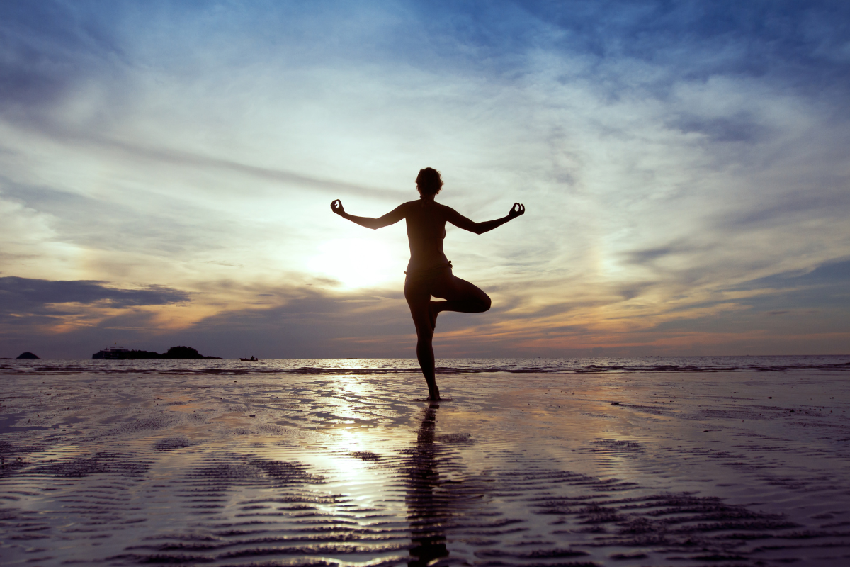 A silhouette image of a person standing on a beach looking towards the ocean with their arms out and balancing on a single leg.