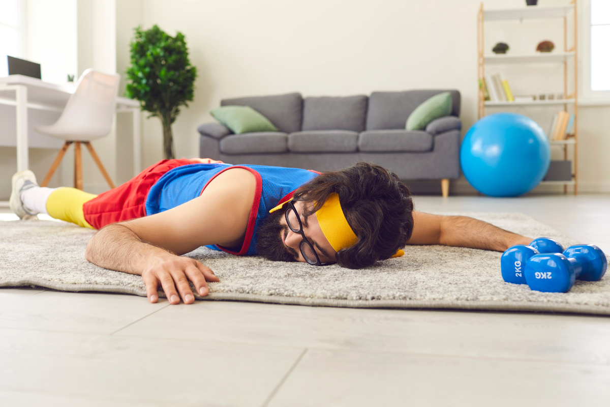 A bearded man lying on the carpet in a living room. He appears to have had a funniest gym fail. He is wearing bright workout clothes and has two small dumbbells on the ground next to him.