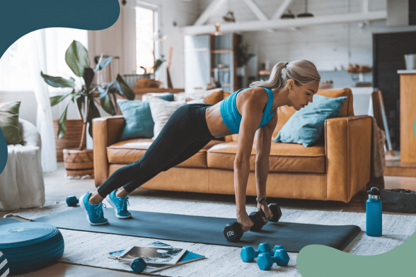 A woman in her living room supporting herself with dumbbells as she does a lower glute workout. She is on a yoga mat in her living room.